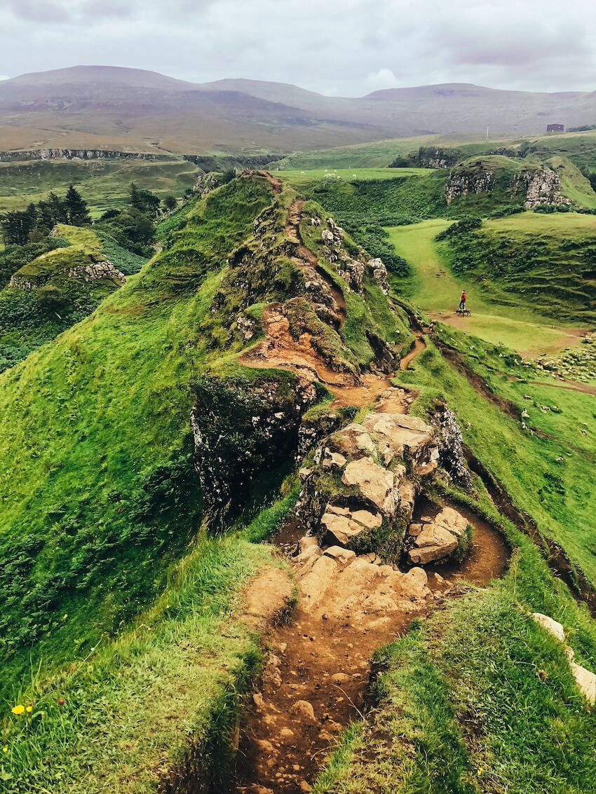 Pohled z úzkého kamenitého hřebene na zelené travnaté kopečky v údolí Fairy Glen na ostrově Skye ve Skotsku. V dálce jsou vidět hory a osamělý turista v roce 2026.