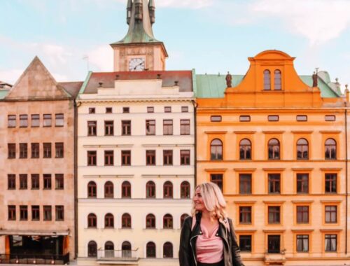 Blonďatá cestovatelka Blond on the road v černé kožené bundě se usmívá a dívá se doprava. V pozadí je ikonické Smetanovo nábřeží v Praze s barevnými historickými domy a věží Staroměstské vodárny u Karlova mostu za jasného dne. Úvodní fotka k článku Co vidět v Praze, Blond on the road.
