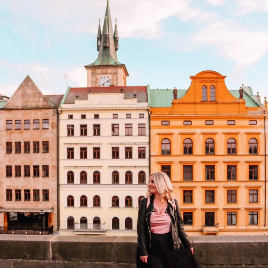 Blonďatá cestovatelka Blond on the road v černé kožené bundě se usmívá a dívá se doprava. V pozadí je ikonické Smetanovo nábřeží v Praze s barevnými historickými domy a věží Staroměstské vodárny u Karlova mostu za jasného dne. Úvodní fotka k článku Co vidět v Praze, Blond on the road.