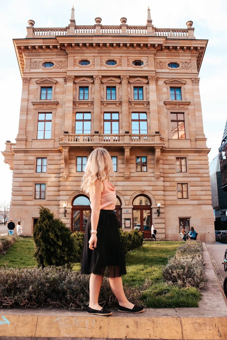 Blonďatá cestovatelka Blond on the road v růžovém topu a černé sukni stojí a dívá se na majestátní fasádu Národního divadla v Praze. Fotka zachycuje boční pohled na budovu s balkony a zlatými detaily na střeše. Fotka v článku Co vidět v Praze.