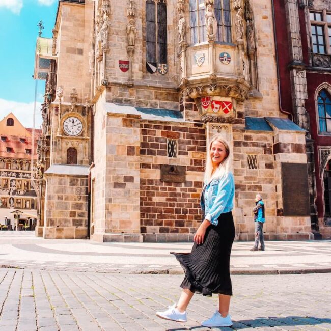 Usměvavá blonďatá cestovatelka Blond on the road v džínové bundě a černé skládané sukni kráčí po dlažbě Staroměstského náměstí v Praze. V pozadí je Staroměstská radnice s detaily orloje a historickou architekturou za slunečného dne. Fotka k článku Co vidět v Praze.