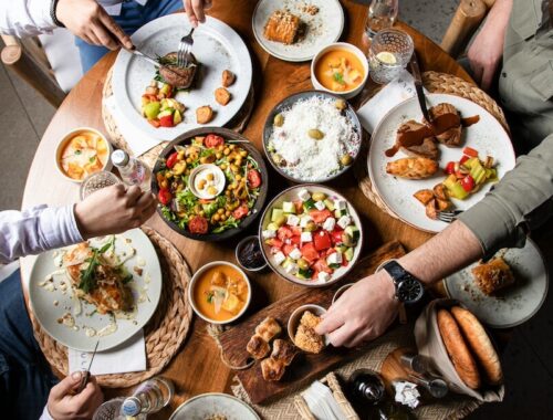 Kam na jídlo v Praze: úvodní fotografie k článku, stůl plný talířů s jídlem a pitím, ruce lidí