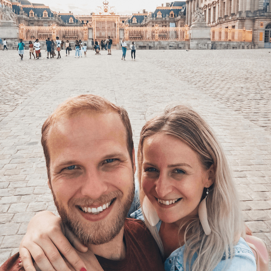 Mladý usmívající se pár stojící před branami paláce ve Versailles. Na fotoografii je autorka blogu Blond on the Road s manželem.