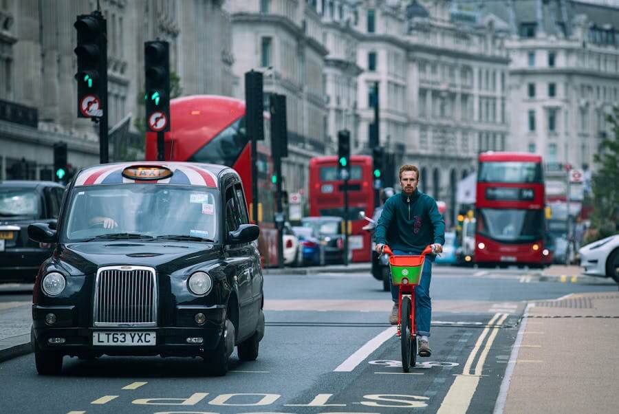 Ranní doprava v Londýně. Tradiční černý londýnský taxík (Black Cab) a cyklista v tyrkysové mikině jedou v pruzích na rušné ulici s červenými doubledeckery v pozadí.