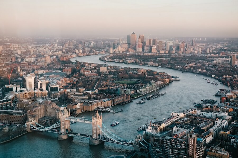 Panoramatický letecký pohled na Tower Bridge a řeku Temži v oparu. Na řece plují lodě, v pozadí mrakodrapy, ilustrující různé formy dopravy v Londýně.
