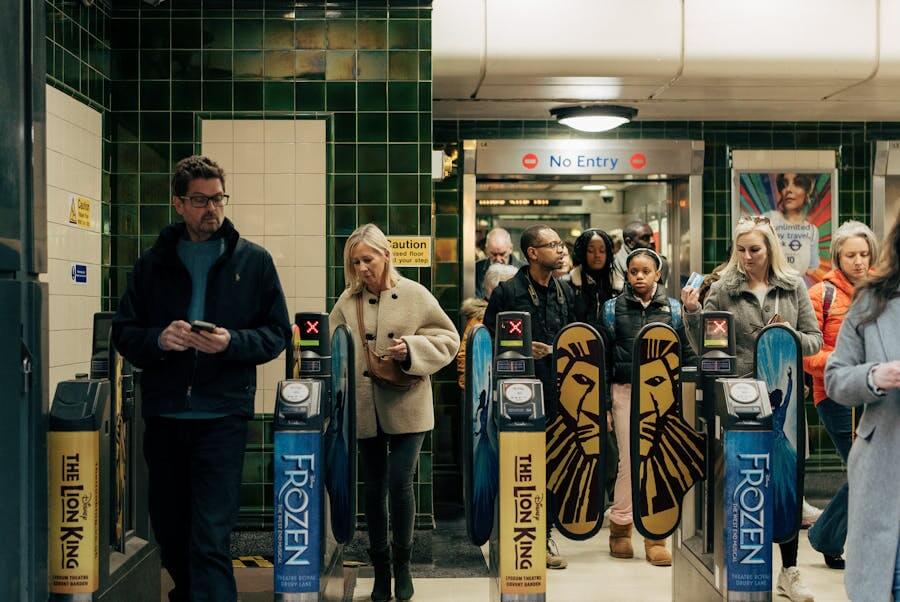 Cestující procházející turnikety v londýnském metru pomocí bezkontaktní platby. Detail na moderní brány s reklamami na muzikály The Lion King a Frozen, ilustrující, jak probíhá doprava v Londýně.