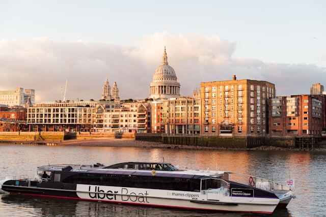 Moderní loď Uber Boat by Thames Clippers plující po řece Temži v Londýně. V pozadí je katedrála svatého Pavla (St Paul's Cathedral) a historické budovy nábřeží při západu slunce, ilustrující alternativní dopravu v Londýně.