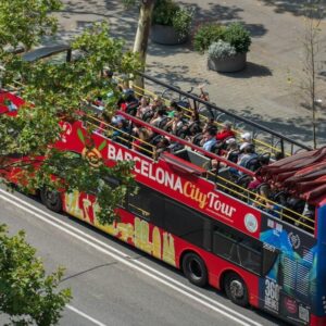 Červený dvoupatrový autobus Barcelona City Tour s turisty na palubě, projíždějící po bulváru v centru města.
