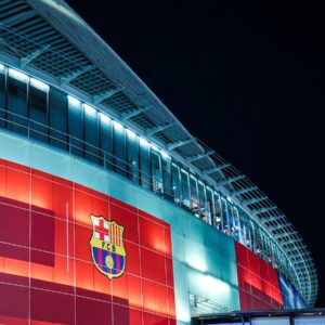 Noční záběr na moderní, osvětlenou fasádu stadionu Camp Nou s velkým logem fotbalového klubu FC Barcelona.