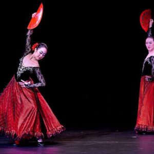 Dvě tanečnice v tradičních červeno-černých šatech tančí flamenco na pódiu a drží vějíře.