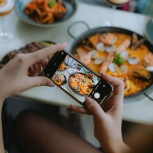 Detailní záběr na mobilní telefon fotící tradiční španělskou paellu s mořskými plody v barcelonské restauraci.