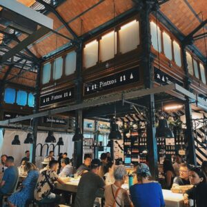 Atmosféra uvnitř tržnice Mercat de la Boqueria, kde lidé sedí u baru a vychutnávají si čerstvé tapas a pintxos Tradiční španělské tapas v tržnici La Boqueria jako chutná součást našeho Barcelona itineráře.