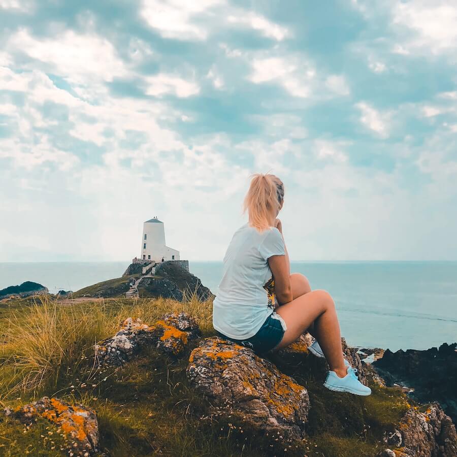 Dívka - autorka blogu Blond on the Road - sedící na kameni na skále, v dálce maják a moře. Nejhezčí místa ve Walesu.