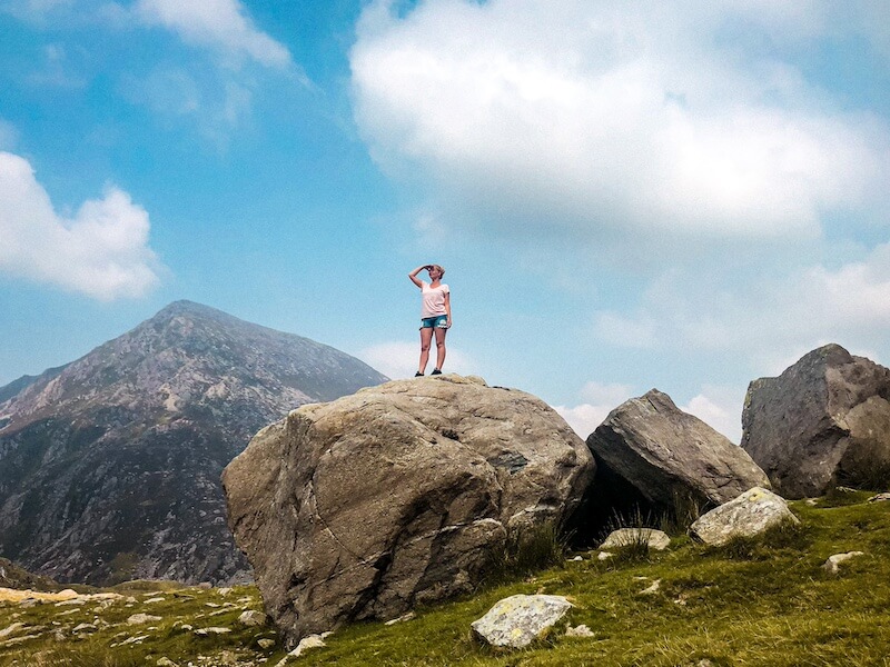 Turistka na skalním balvanu s horou Tryfan v pozadí, Eryri, Wales