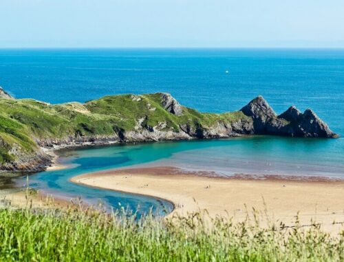 Pohled na pláž Three Cliffs Bay s tyrkysovým mořem a skalnatými výběžky. Místa ve Walesu, která vypadají jako Nový Zéland
