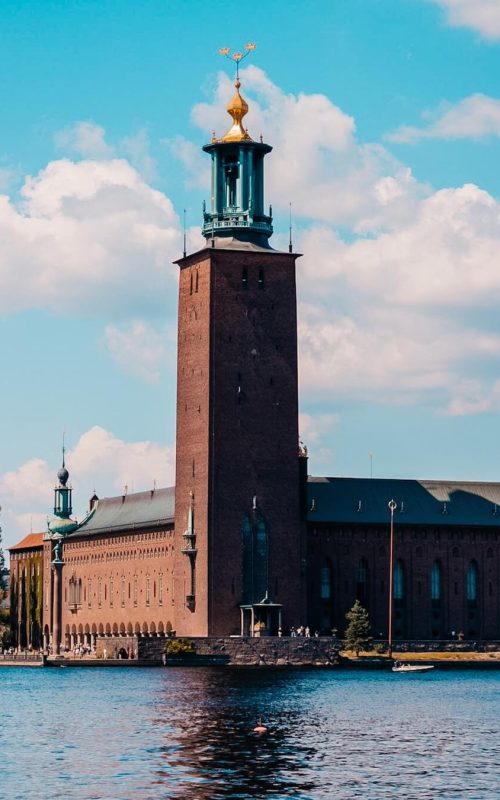 Ikonická věž Radnice Stadshuset jako hlavní bod toho, co vidět ve Stockholmu. Pohled z vody na cihlovou architekturu se zlatými korunkami během slunečného dne.