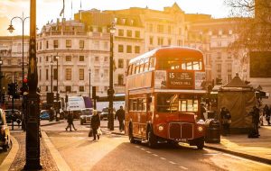 Romantický záběr na historický červený autobus Routemaster na lince 15 směrem na Tower Hill, projíždějící zalitou ulicí v centru. Závěrečný tip pro romantickou dopravu v Londýně při západu slunce.