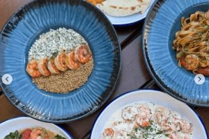 Bohaté plody moře v restauraci Černomorka v Praze: Pohled shora na stůl s různými talíři plnými krevet, těstovin a omáček v modrých miskách. Skvělý tip pro milovníky seafoodu při hledání, kam na jídlo v Praze.