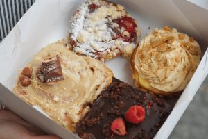 Nejlepší cukrárny v Praze: Pohled do papírového boxu na čtyři různé, bohatě zdobené donuty (Kinder Bueno, malinový, čokoládový, skořicový). Sladké gastro zážitky v Praze v podniku Kus Koláče.