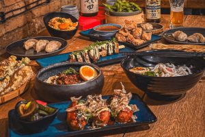 Stůl plný asijských specialit v Praze: Sushi sety, gyoza knedlíčky, edamame boby, sake a japonské pivo na dřevěném stole. Inspirace pro nejlepší asijskou restauraci v Praze - Teplá Kačka.