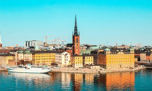 Slunečné panorama Stockholmu z výšky s výhledem na barevné historické budovy, moře a výraznou centralní kostelní věž. Úvodní fotka k článku Stockholm levně.