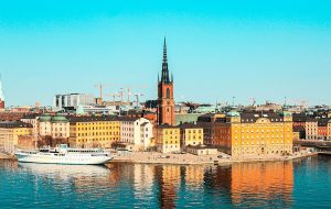 Slunečné panorama Stockholmu z výšky s výhledem na barevné historické budovy, moře a výraznou centralní kostelní věž. Úvodní fotka k článku Stockholm levně.