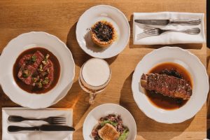 Fotografie focená shora pohled na 4 talíře české kuchyně, maso, ryby a salát, příbory po stranách talířů. Jídlo z restaurace Kuchyň na Pražském hradě v Praze.