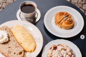 Bohatý venkovní brunch v podniku Moe v Holešovicích na modrém stole u dlážděné ulice: talíř s nadýchanou vaječnou omeletou a kváskovým chlebem, miska s müsli a ovocem, sladký skořicový šnek a šálek černé kávy. Ideální místo pro ranní kávu při plánování, kam na jídlo v Praze.