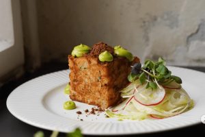 Detailně aranžovaná velikonoční nádivka ve tvaru kostky, zdobená kapkami avokádového krému a bylinkami, podávaný s ředkvičkovým salátem. Ukázka moderní gastronomie, kterou můžete ochutnat, když hledáte, kam na jídlo v Praze na brunch. Podnik Blumery v Praze na Vinohradech.