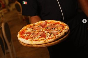 Nejlepší pizza v Praze - Da Pietro Praha: Kuchař v rondonu s italskou vlajkou drží čerstvě upečenou pizzu s parmskou šunkou a žampiony na dřevěném prkénku. Gastro zážitek v centru Prahy.