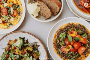 Pestrobarevná nabídka vegetariánských pokrmů shora na šedém stole: miska čočkového salátu s rajčaty a bylinkami, pečená zelenina a košíček s kváskovým chlebem a máslem. Skvělý tip, kam na jídlo v Praze na zdravý oběd nebo brunch, Etapa Karlín.