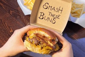 Burger z restaurace FatFuck Smash Burger v Praze v papirové krabičce, burger držený v rukou