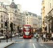 Doprava v Londýně: Pohled na rušnou Regent Street s ikonickým červeným patrovým autobusem (Doubledeckerem), typickými černými taxíky a historickými budovami v pozadí.